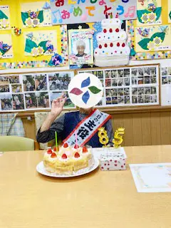 グループホーム朝風 誕生日会。ろうそくを立てたケーキを前にたすきをかけてお祝いされる入居者